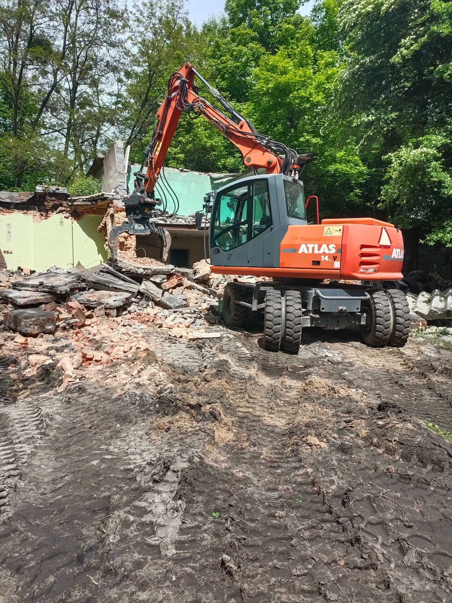 Pomarańczowy koparko-ładowarka Atlas 1404 V z chwytakiem hydraulicznym kruszy resztki budynku z zielonymi ścianami, widoczne gruz i ślady opon na błotnistym gruncie, drzewa w tle.