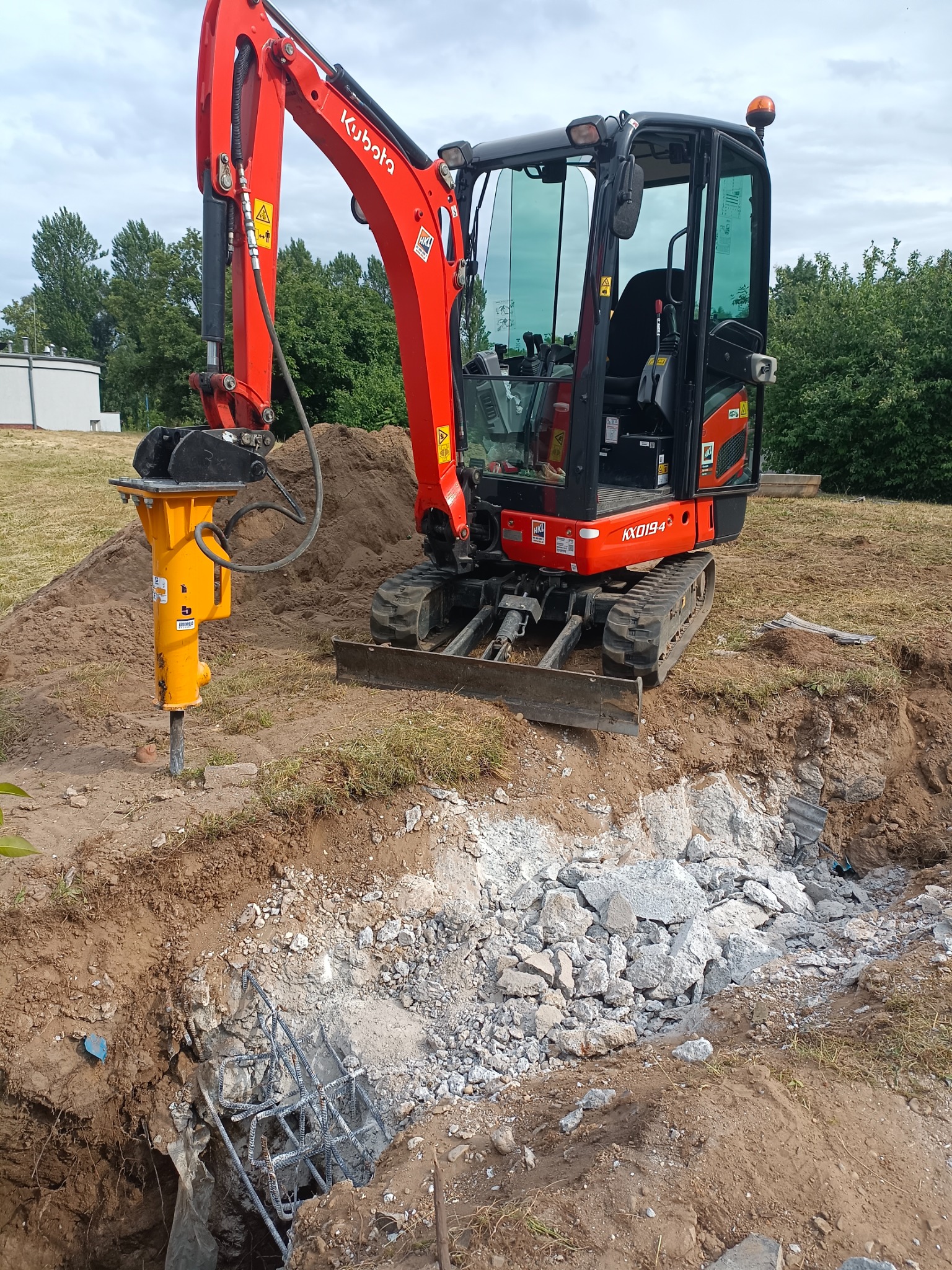 Czerwona minikoparka Kubota z żółtym młotem hydraulicznym rozbija betonowe elementy w wykopie ziemnym z widocznym zbrojeniem.
