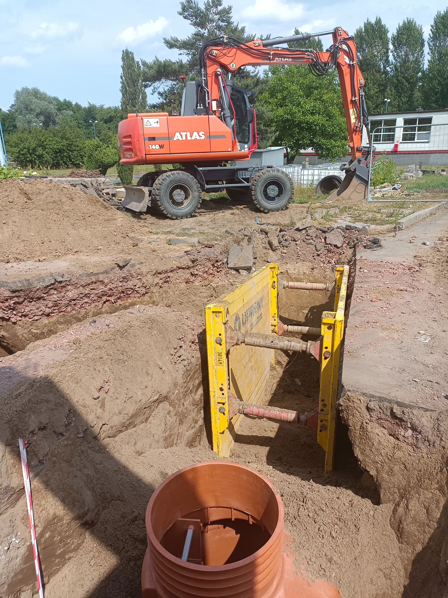 Pomarańczowa koparka Atlas 140W na placu budowy przy wykopie zabezpieczonym żółtymi szalunkami, widoczny element instalacji kanalizacyjnej z tworzywa sztucznego.