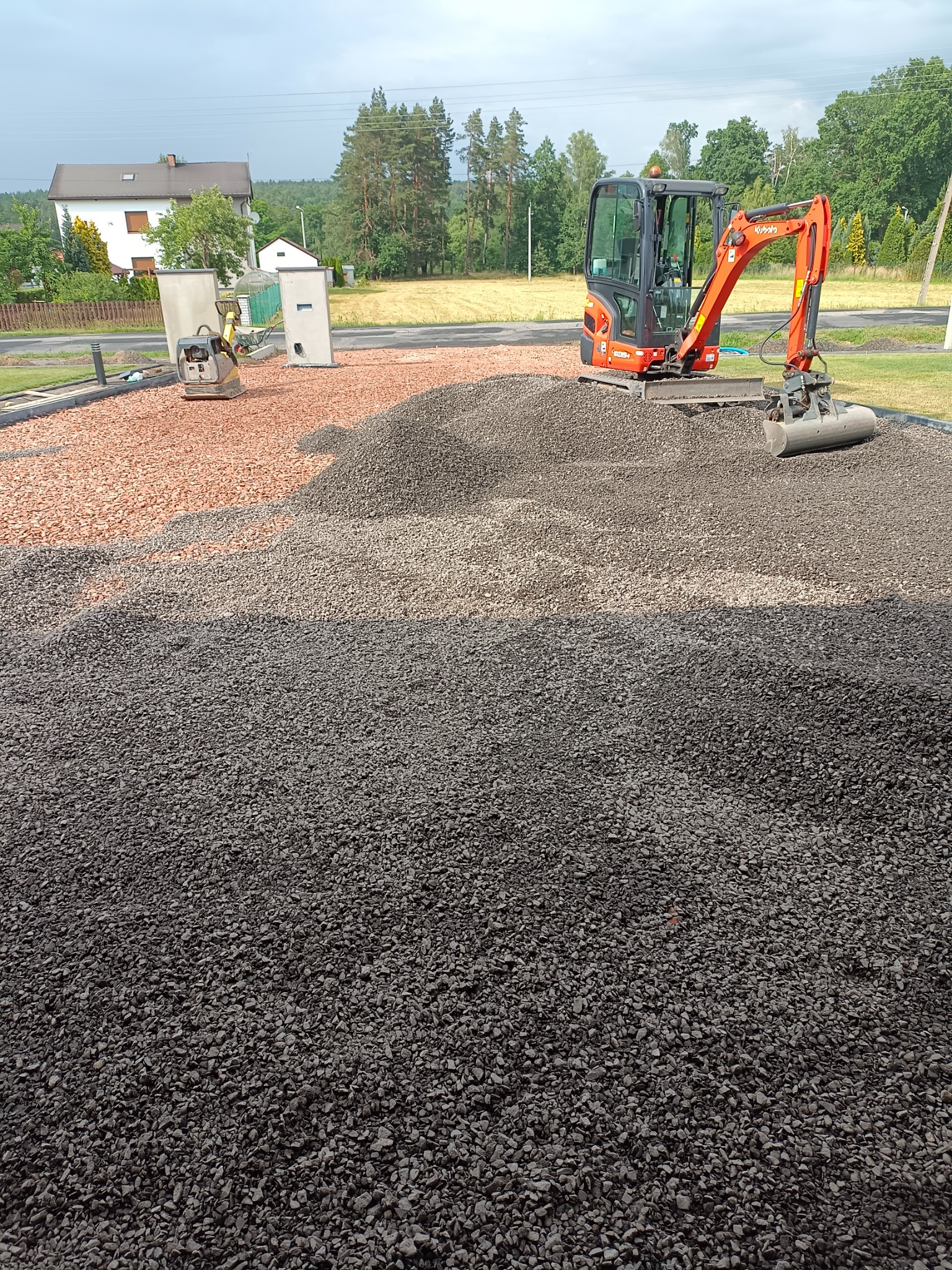 Pomarańczowa mini koparka Kubota na stercie ciemnego gruzu, w tle czerwony gruz, zagęszczarka i domy jednorodzinne.