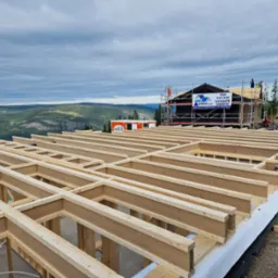 Wznoszenie konstrukcji drewnianej budynku w malowniczym górskim krajobrazie, widoczny szkielet parteru z belek, rusztowania przy budynku w tle, tablica informacyjna na elewacji.