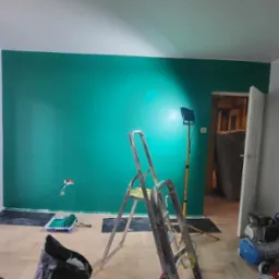 Świeżo pomalowana ściana w odcieniach zieleni, oświetlona lampą roboczą, z widocznymi narzędziami malarskimi, takimi jak drabina, kuweta z wałkiem i kompresor, na podłodze zabezpieczonej folią.