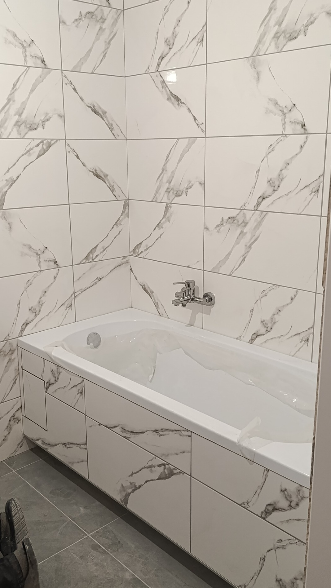 Wykończona wanna obudowana płytkami imitującymi marmur, z baterią ścienną, otoczona ścianami wyłożonymi tymi samymi płytkami, szare płytki podłogowe.