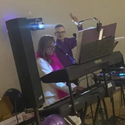 Dwoje muzyków grających na keyboardach Yamaha i Korg podczas występu, kobieta z różowym boa na szyi, mężczyzna w fioletowej koszuli z mikrofonem w dłoni, oświetlenie sceniczne i balony w tle.