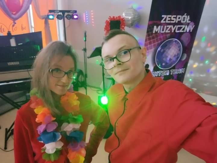 Dwoje muzyków w czerwonych koszulach pozuje do selfie przed sceną z klawiszami Yamaha, kolorowymi światłami i banerem z napisem 'Zespół Muzyczny'. Kobieta ma na szyi hawajski naszyjnik z kwiatów.