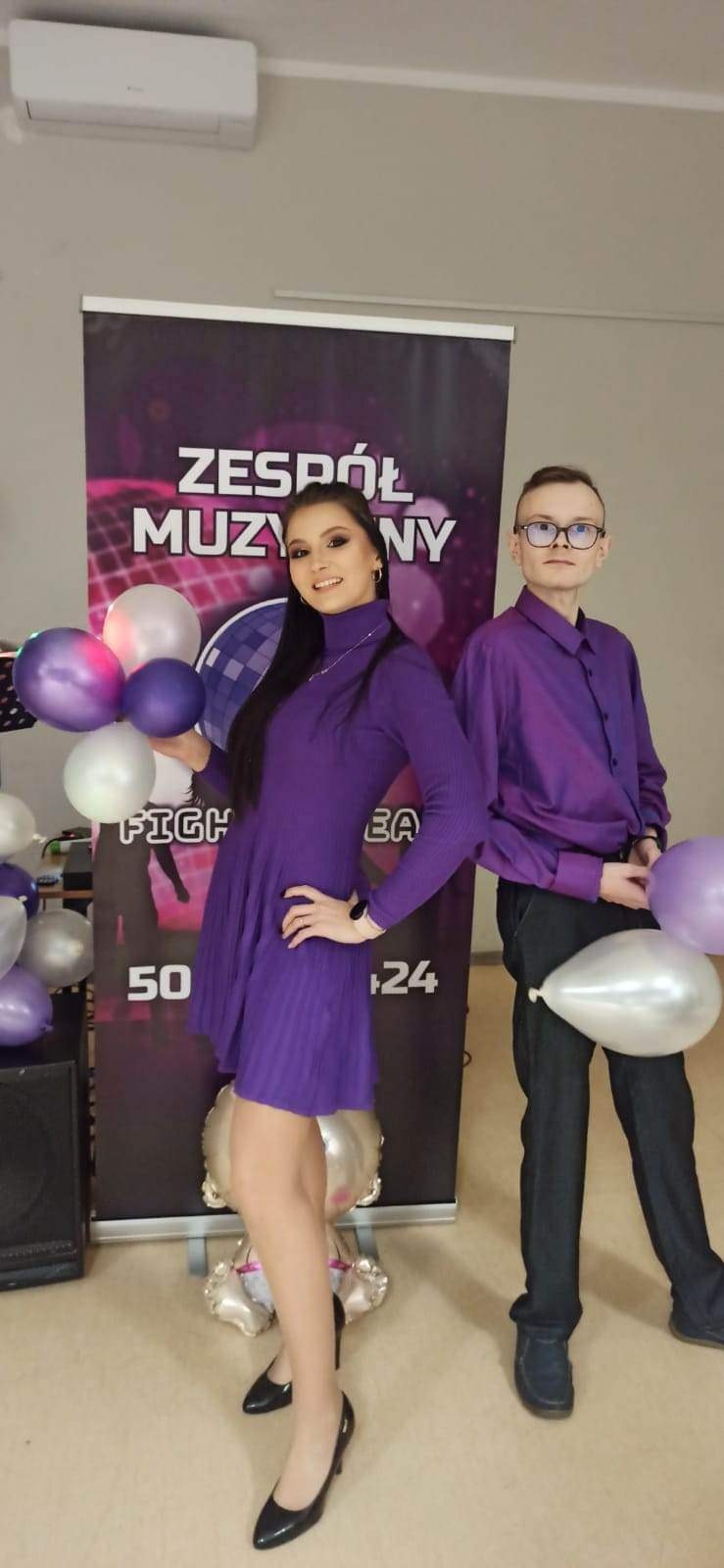 Dwoje młodych ludzi w fioletowych strojach pozuje na tle baneru z napisem 'Zespół Muzyczny', trzymając fioletowe i białe balony.