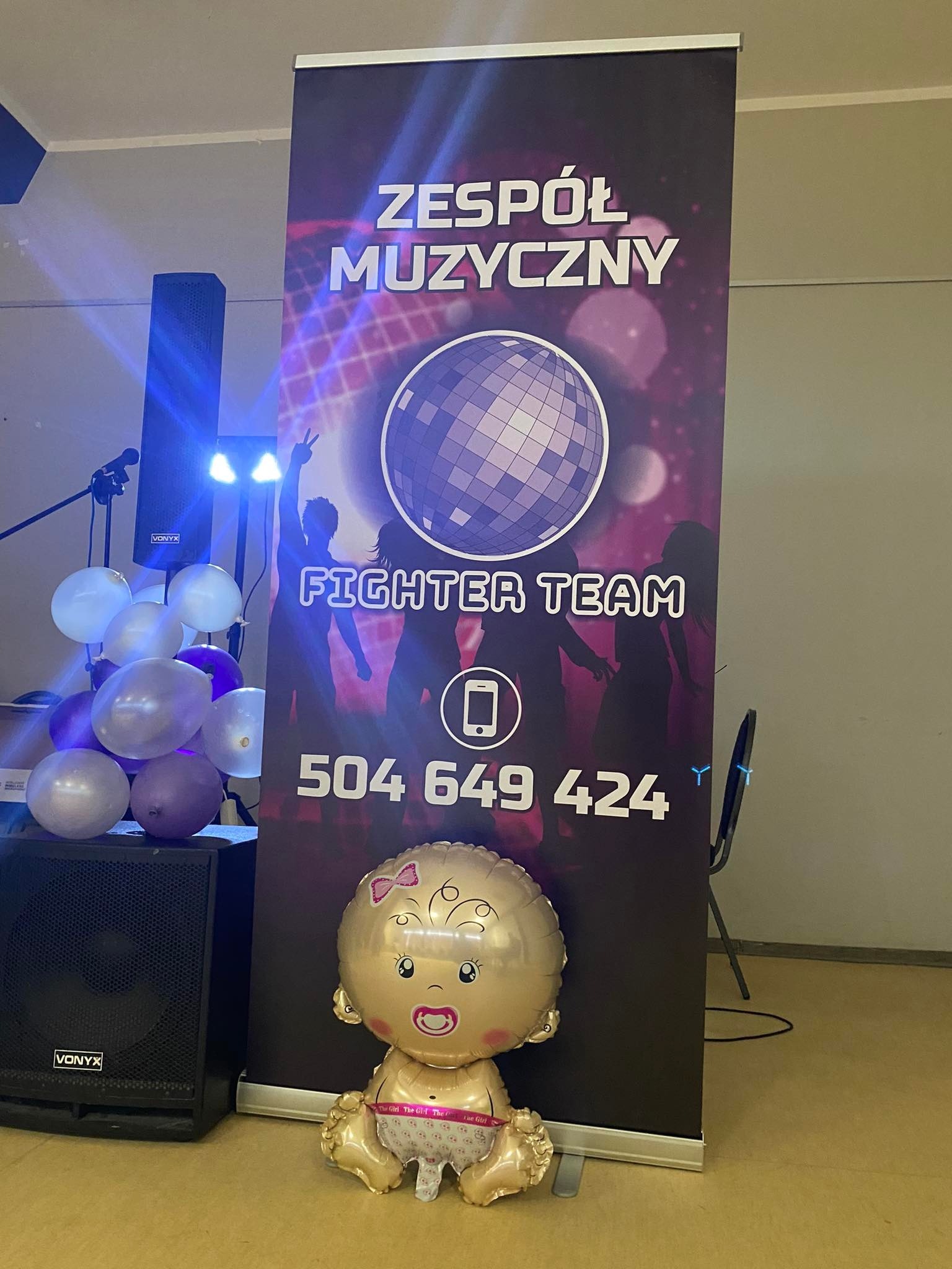Roll-up reklamowy z nazwą 'Zespół Muzyczny Fighter Team' i numerem telefonu, obok głośnik marki Vonyx, balony i foliowy balon w kształcie niemowlaka.