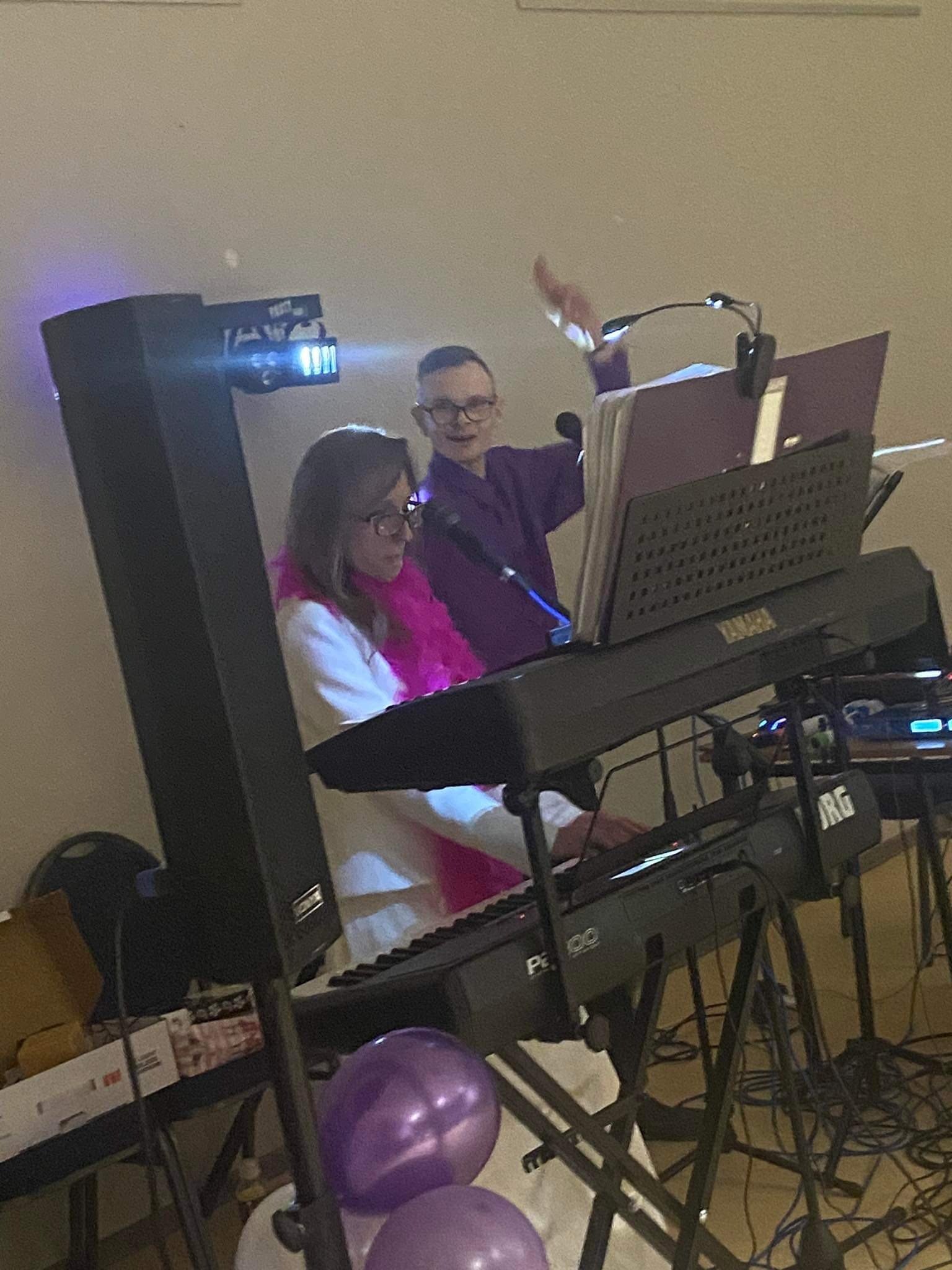 Dwoje muzyków grających na keyboardach Yamaha i Korg podczas występu, kobieta z różowym boa na szyi, mężczyzna w fioletowej koszuli z mikrofonem w dłoni, oświetlenie sceniczne i balony w tle.