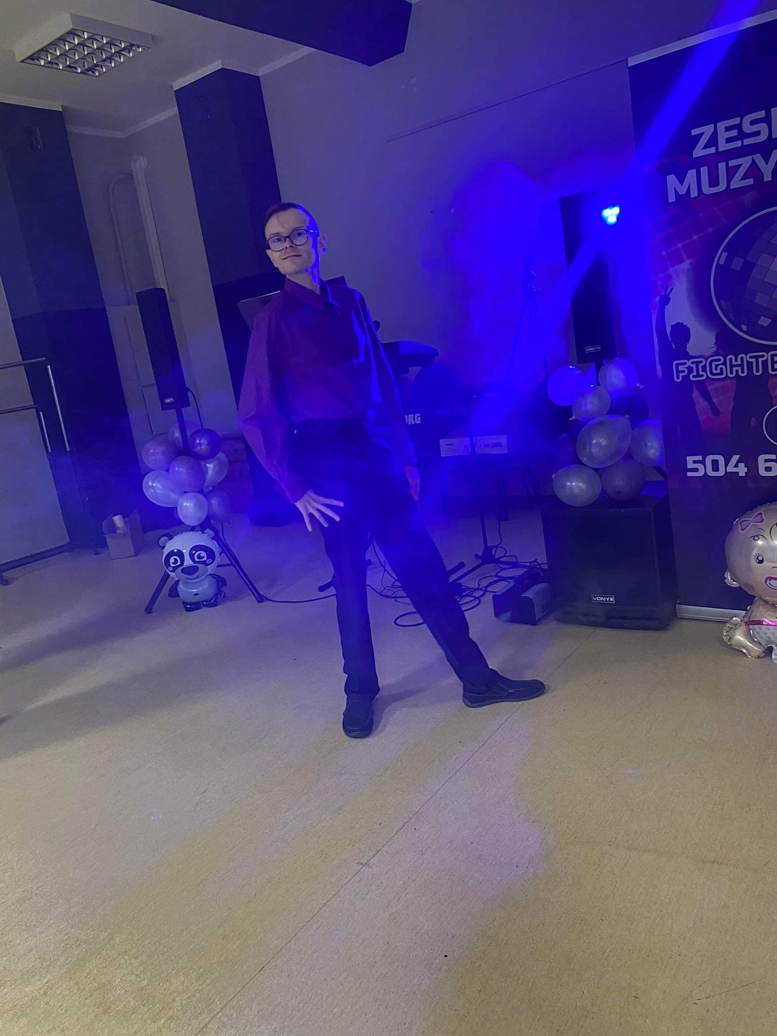 Mężczyzna w okularach i bordowej koszuli pozuje na tle sceny z balonami, nagłośnieniem i plakatem zespołu muzycznego z efektem niebieskich świateł.