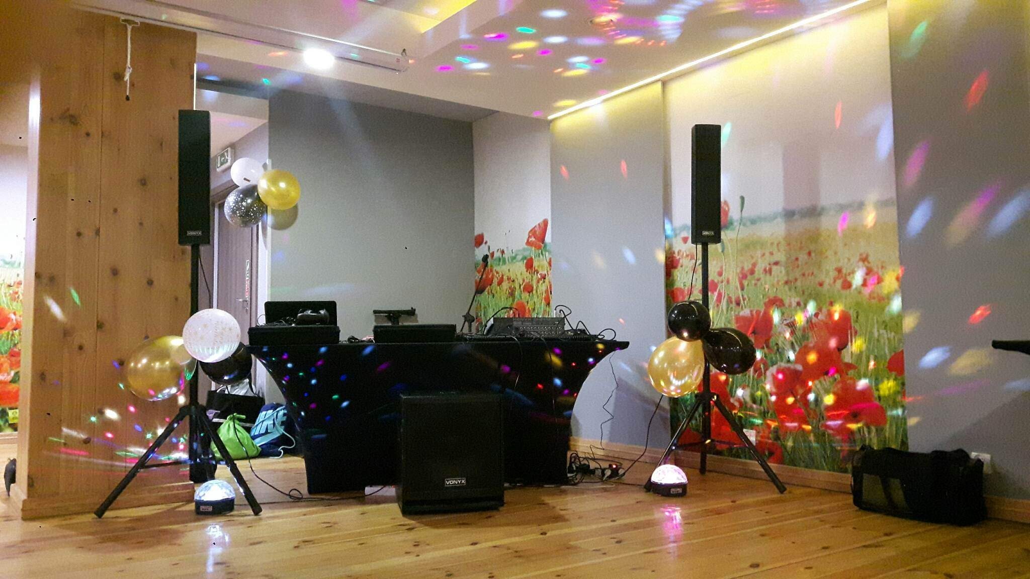 Sprzęt DJ-ski: konsoleta pokryta czarną tkaniną z kolorowymi światłami, głośniki na statywach, kula dyskotekowa i balony w sali z drewnianą podłogą i ścianą z motywem polnych maków.