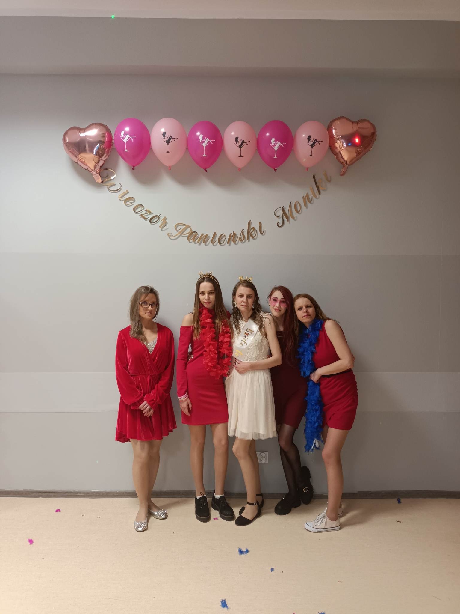 Pięć kobiet pozuje do zdjęcia na tle ściany z balonami i napisem 'Wieczór Panieński Monki', ubrane w sukienki i boa, jedna z szarfą 'Bride to be'.