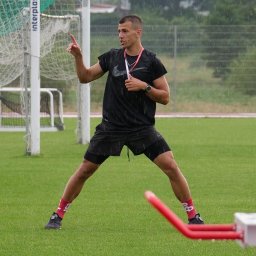 GALIKTEAM Michał Galikowski - Młody mężczyzna w sportowym stroju, gestykulujący na mokrym boisku podczas deszczu, w tle siatka do piłki nożnej.