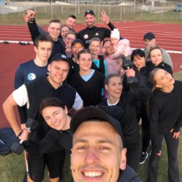 GALIKTEAM Michał Galikowski - Selfie grupy uśmiechniętych osób z hantlami na bieżni stadionu, w tle budynki i drzewa.