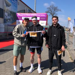 GALIKTEAM Michał Galikowski - Trzech mężczyzn pozuje do zdjęcia po zawodach duathlonowych, widoczne numery startowe 393 i 392, butelki z wodą i budynek z reklamą w tle.