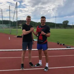 Dwóch uśmiechniętych mężczyzn ściska sobie dłonie na czerwonej bieżni stadionu, w tle leżąca osoba, butelka wody, kettlebells i mata do ćwiczeń.