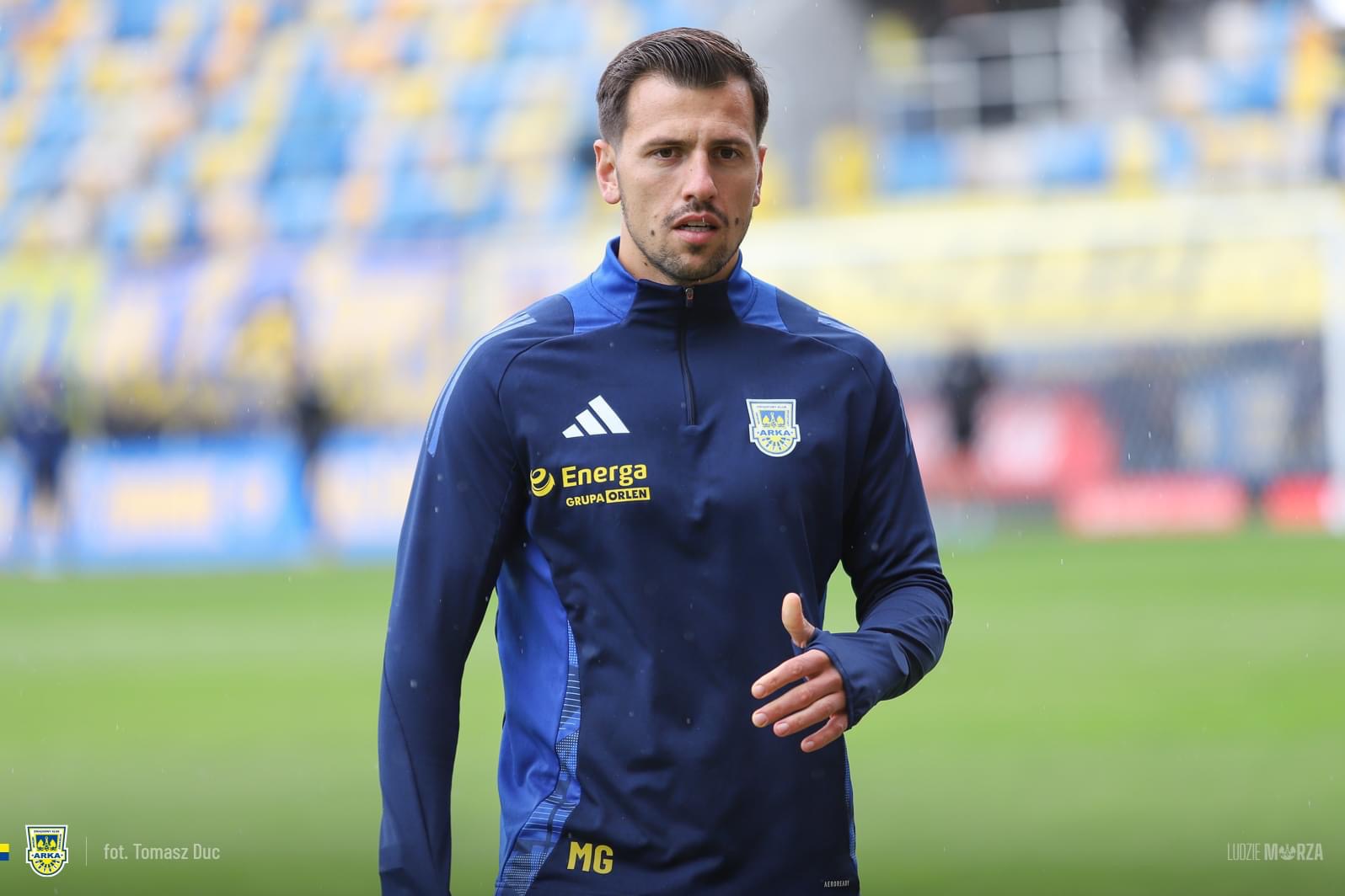 Mężczyzna w sportowym stroju z logotypami sponsorów, stojący na tle stadionu.