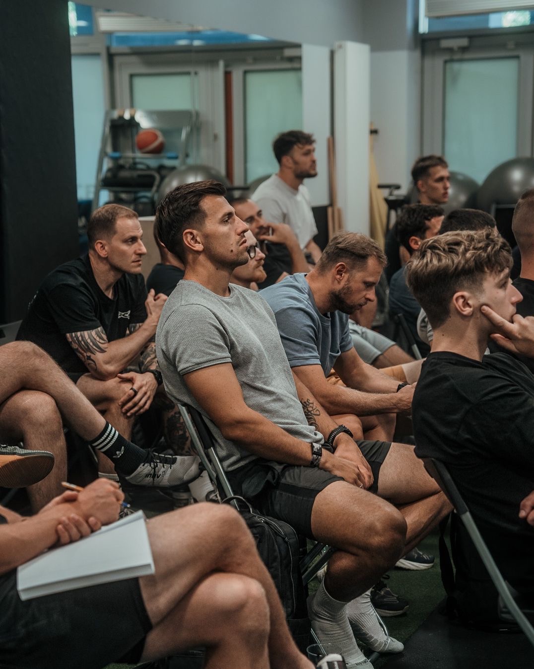 Grupa mężczyzn siedzi na krzesłach w sali gimnastycznej, słuchając prelekcji, z notatnikiem i długopisem w rękach, widoczne piłki do ćwiczeń w tle.