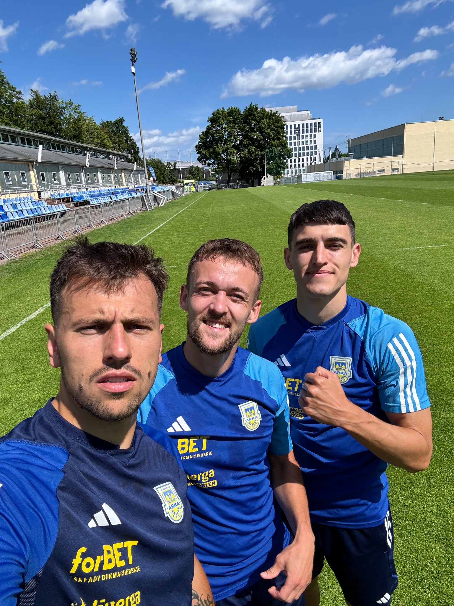 Trzech młodych mężczyzn w niebieskich strojach sportowych z logo drużyny piłkarskiej na tle boiska i trybun stadionu w słoneczny dzień.