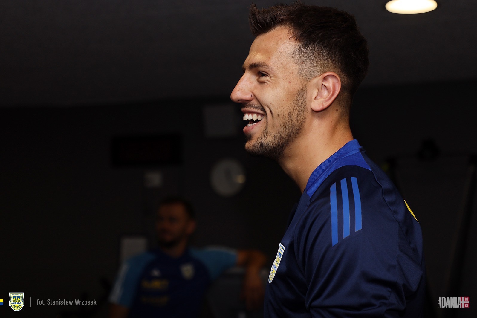 Uśmiechnięty mężczyzna z krótką brodą, ubrany w granatową koszulkę sportową z niebieskimi paskami na ramieniu, patrzy w prawo; w tle niewyraźna postać.