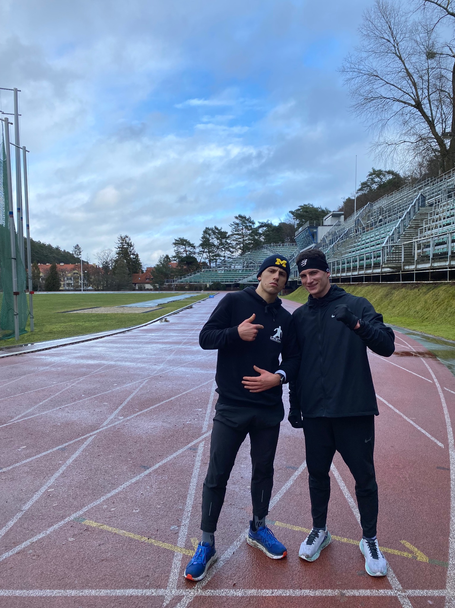 Dwóch młodych mężczyzn na mokrej bieżni stadionu, ubranych w sportowe stroje i czapki, jeden w czapce z daszkiem z literą 'M', drugi w opasce na głowie, w tle trybuny i drzewa.