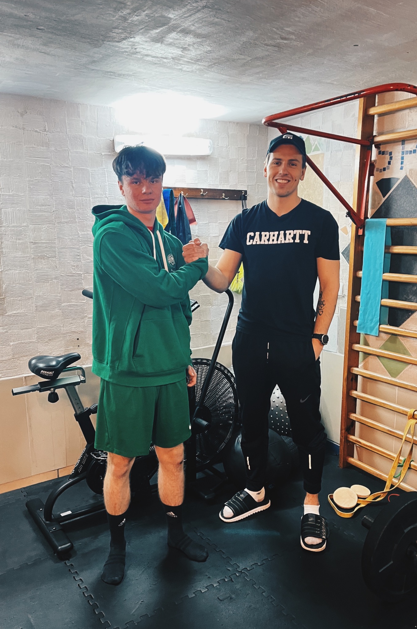 Uśmiechnięty trener w czarnej koszulce Carhartt wita się z klientem w zielonym dresie na tle siłowni z drabinkami, rowerem stacjonarnym i obciążeniami.