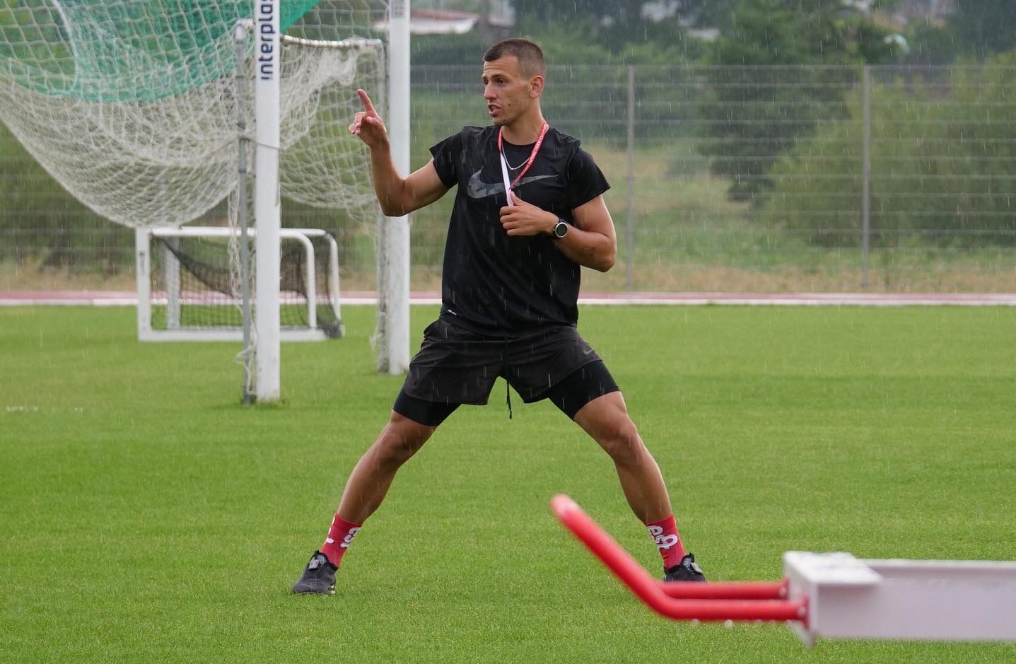 Młody mężczyzna w sportowym stroju, gestykulujący na mokrym boisku podczas deszczu, w tle siatka do piłki nożnej.