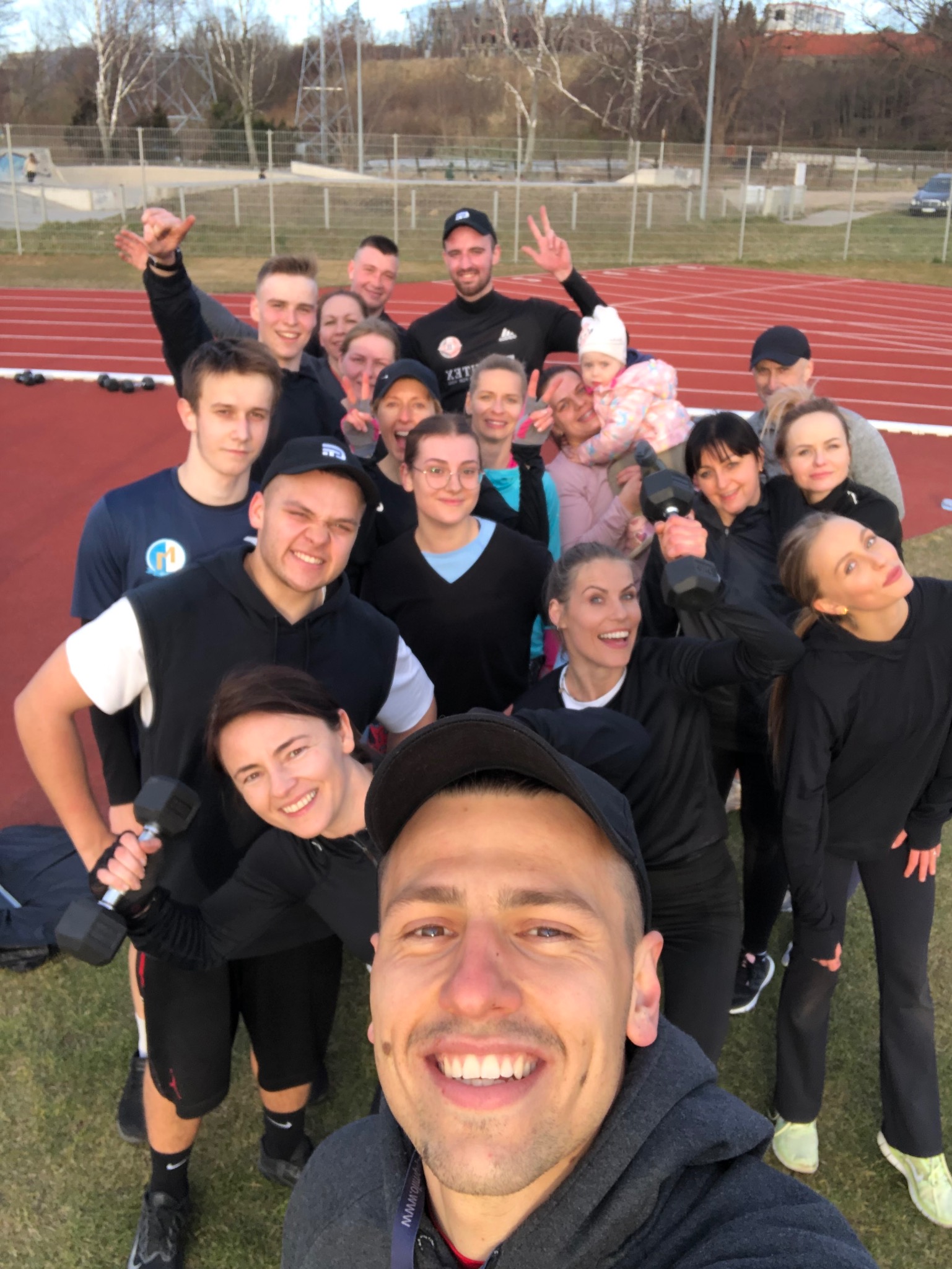 Selfie grupy uśmiechniętych osób z hantlami na bieżni stadionu, w tle budynki i drzewa.