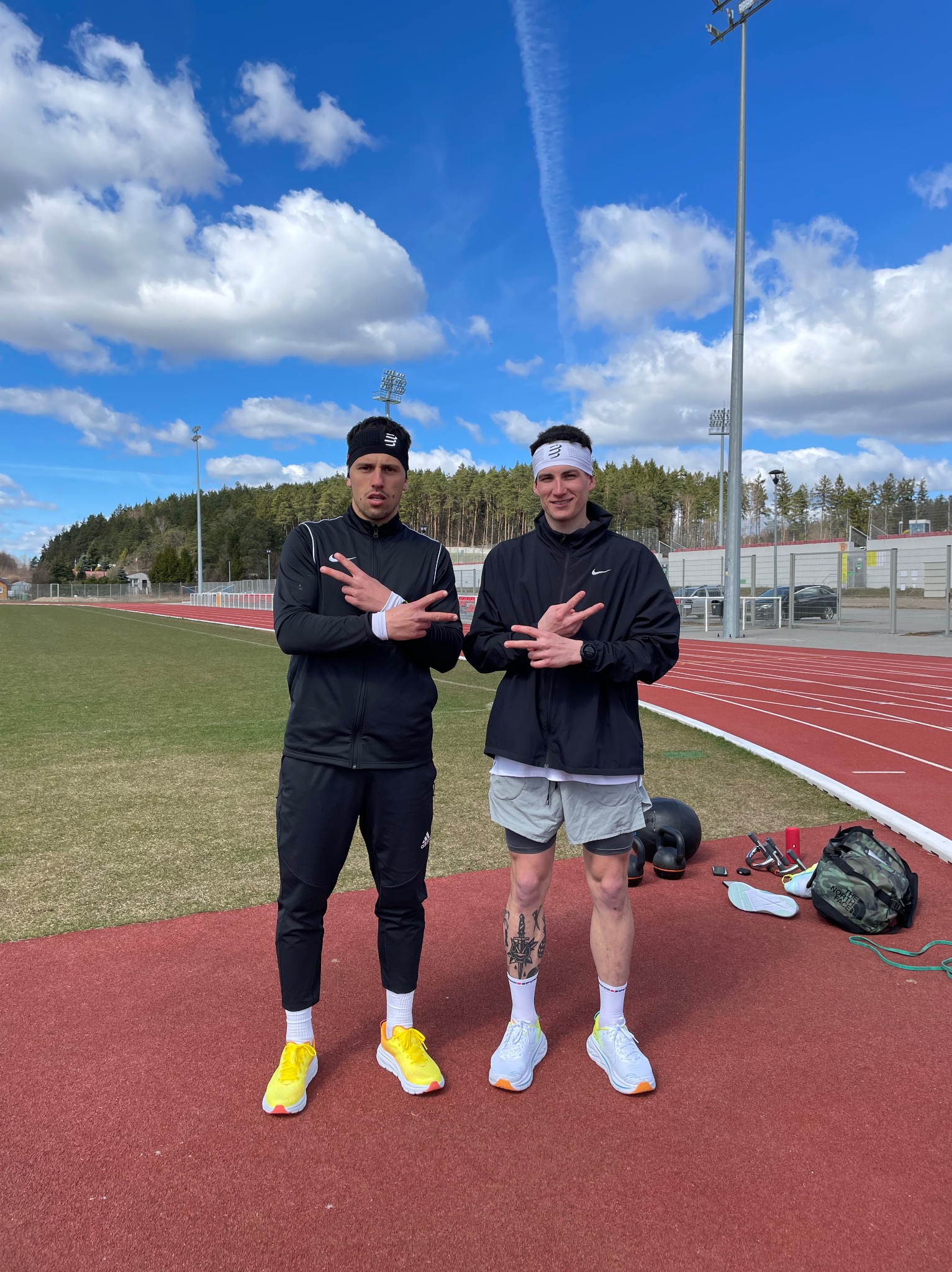 Dwaj mężczyźni na bieżni lekkoatletycznej, ubrani w sportowe stroje i opaski na głowach, pokazują gest zwycięstwa, obok widoczne akcesoria treningowe, w tle zielona trawa i drzewa.