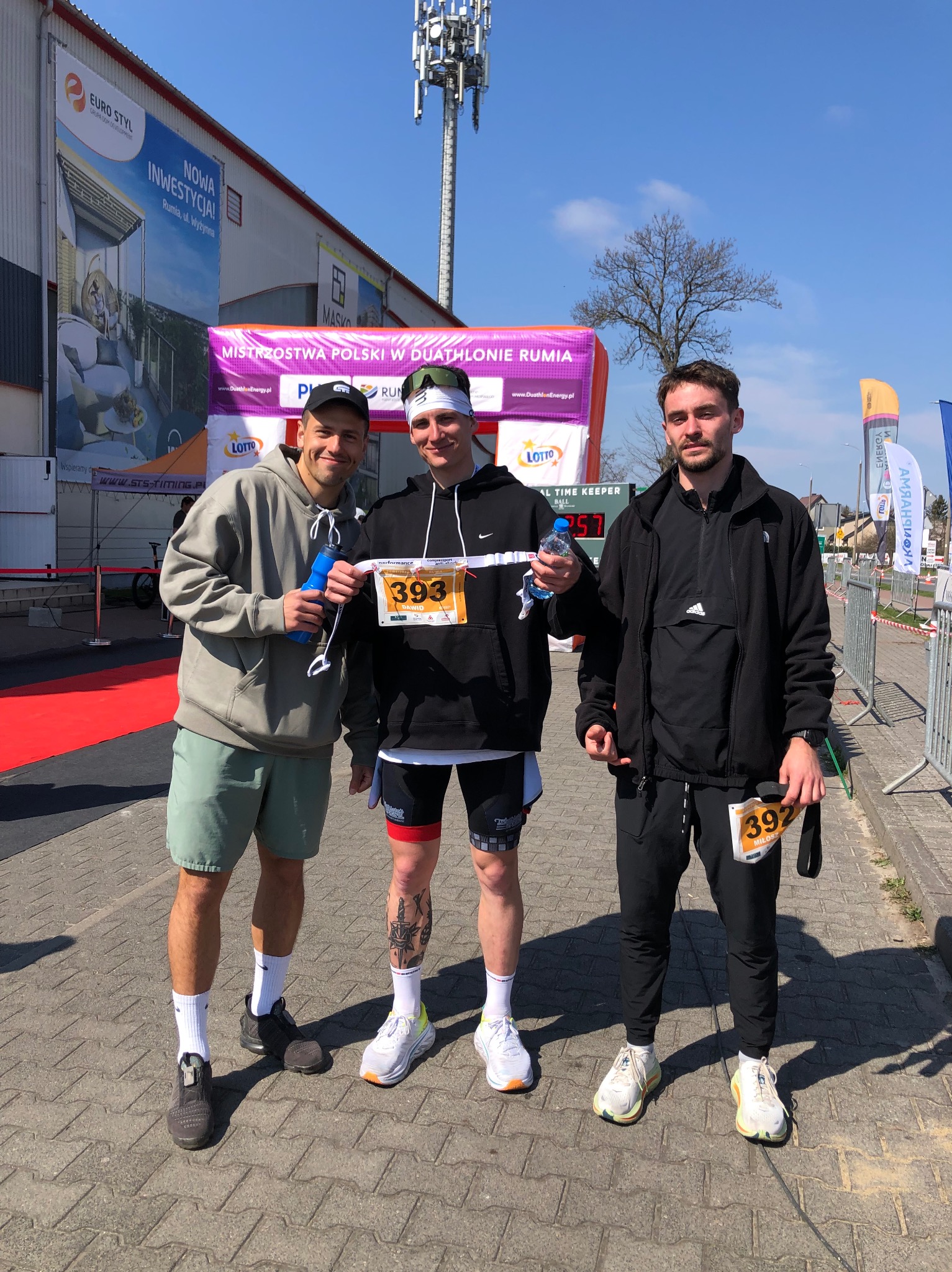 Trzech mężczyzn pozuje do zdjęcia po zawodach duathlonowych, widoczne numery startowe 393 i 392, butelki z wodą i budynek z reklamą w tle.