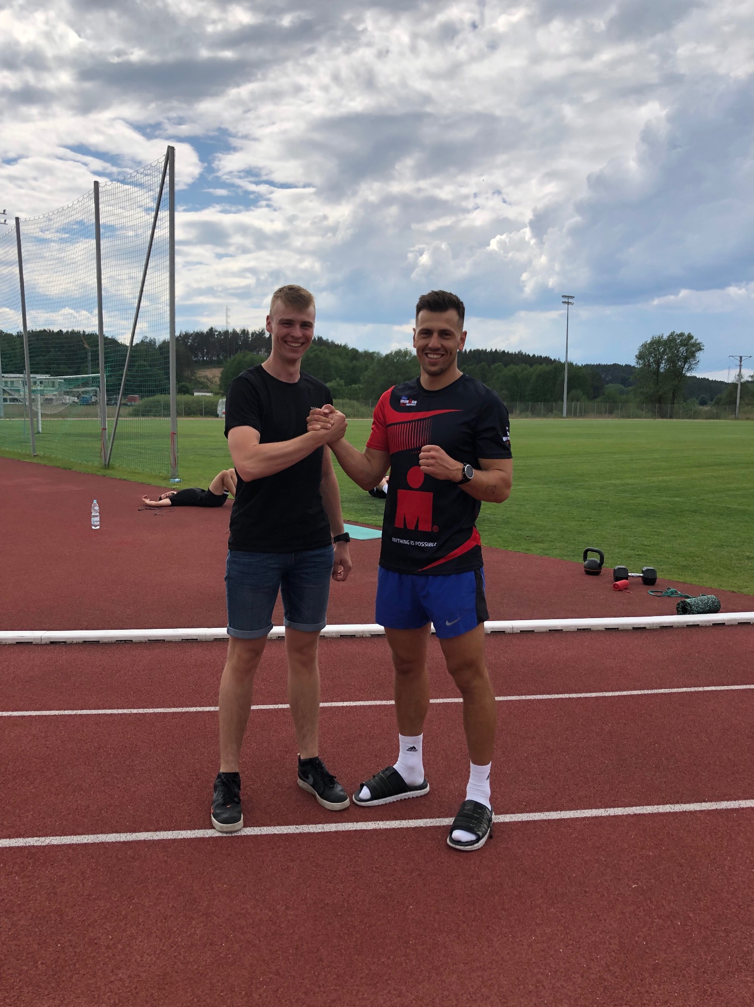 Dwóch uśmiechniętych mężczyzn ściska sobie dłonie na czerwonej bieżni stadionu, w tle leżąca osoba, butelka wody, kettlebells i mata do ćwiczeń.