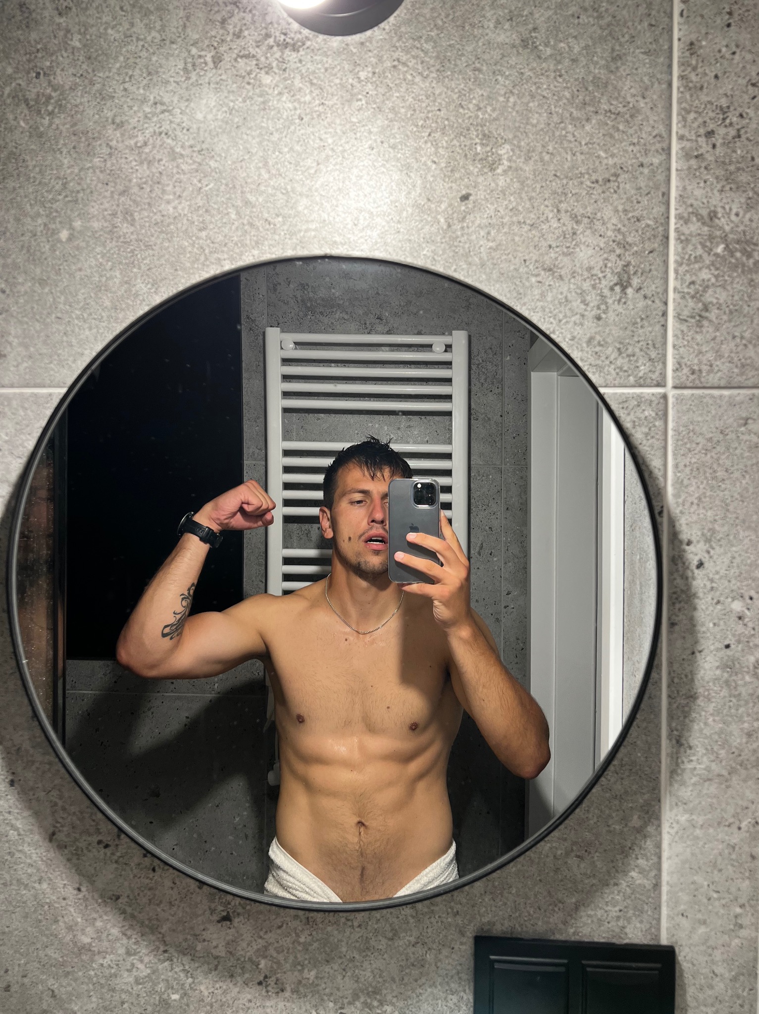 Mężczyzna z widoczną muskulaturą pozuje do selfie w łazience, lekko napinając biceps, z widocznym tatuażem na ramieniu i ręcznikiem w pasie.