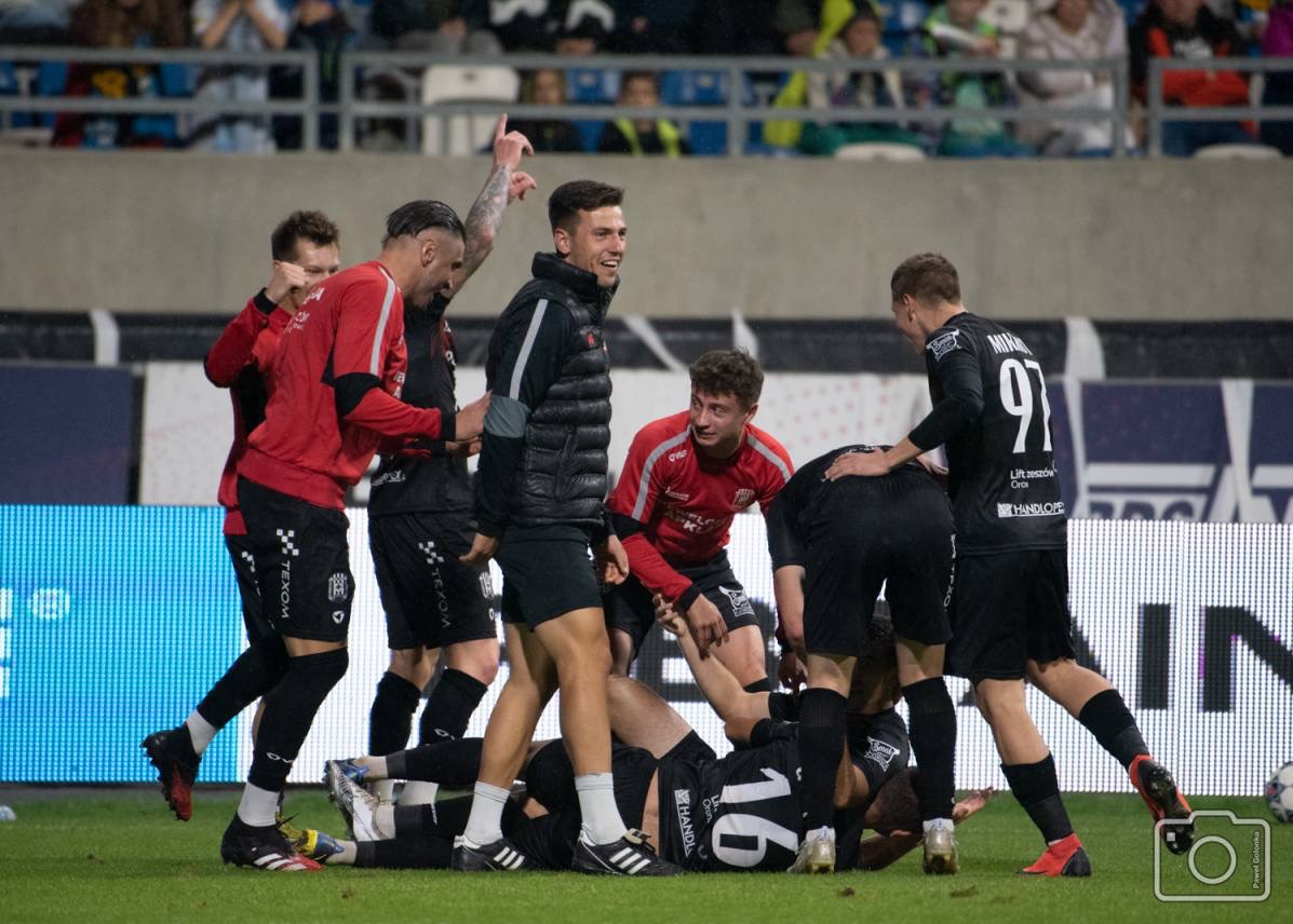 Grupa piłkarzy świętuje zwycięstwo, jeden z nich leży na ziemi, a pozostali stoją wokół niego, w tle widoczni kibice na trybunach.