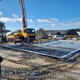 Wylewanie betonu na przygotowany fundament pod budowę domu jednorodzinnego, widoczny mikser do betonu i pompa do betonu na tle zabudowań gospodarczych w zimowej scenerii.
