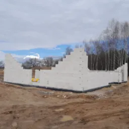 Wznoszenie ścian parterowego domu z białych bloczków silikatowych, widoczne zbrojenie fundamentów, w tle drzewa bez liści i pochmurne niebo.