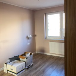 Marcin Pawluczuk Remonty - Pokój po malowaniu ścian na pudrowy róż, z nową podłogą z paneli, widoczny grzejnik pod oknem i kartony na podłodze.