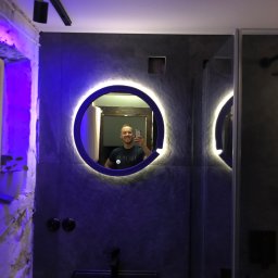 Marcin Pawluczuk Remonty - Nowoczesna łazienka z szarymi płytkami, okrągłym podświetlanym lustrem, czarną umywalką i kabiną prysznicową; odbicie mężczyzny robiącego zdjęcie telefonem.