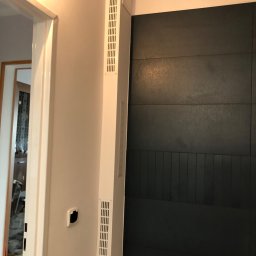 Marcin Pawluczuk Remonty - Fragment ściany z ciemnymi, poziomymi panelami dekoracyjnymi i pionowymi listwami w dolnej części, sąsiadującej z białym narożnikiem z kratkami wentylacyjnymi i otwartym przejściem do innego...