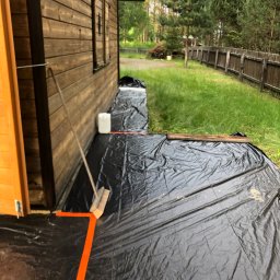 Marcin Pawluczuk Remonty - Przygotowanie do malowania drewnianej elewacji domu: zabezpieczona folią przestrzeń wokół ściany, widoczna szczotka i kanister z farbą.