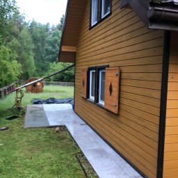 Marcin Pawluczuk Remonty - Elewacja domu z drewnianą okładziną w kolorze miodowym, z brązowymi ramami okiennymi i okiennicami z wyciętym rombem, oraz szarym chodnikiem z płyt betonowych.