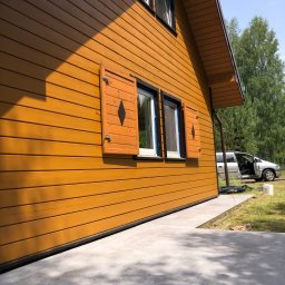 Marcin Pawluczuk Remonty - Żółta, drewniana elewacja domu z brązowymi okiennicami z diamentowym wycięciem, okna zabezpieczone taśmą malarską w Warszawie, widoczny samochód z otwartymi drzwiami.