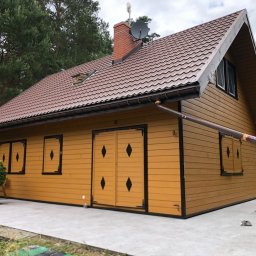 Marcin Pawluczuk Remonty - Żółty, drewniany dom z brązowym dachem z blachy trapezowej, ceglanym kominem i anteną satelitarną, widoczny z zewnątrz na tle drzew.