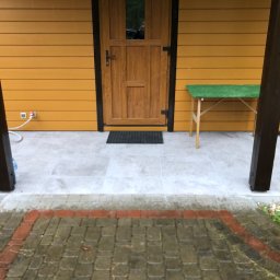 Marcin Pawluczuk Remonty - Wejście do domu z drewnianą elewacją i nową, jasnoszarą nawierzchnią z płytek, widoczne brązowe słupy i stolik z zielonym blatem, kostka brukowa na podjeździe.