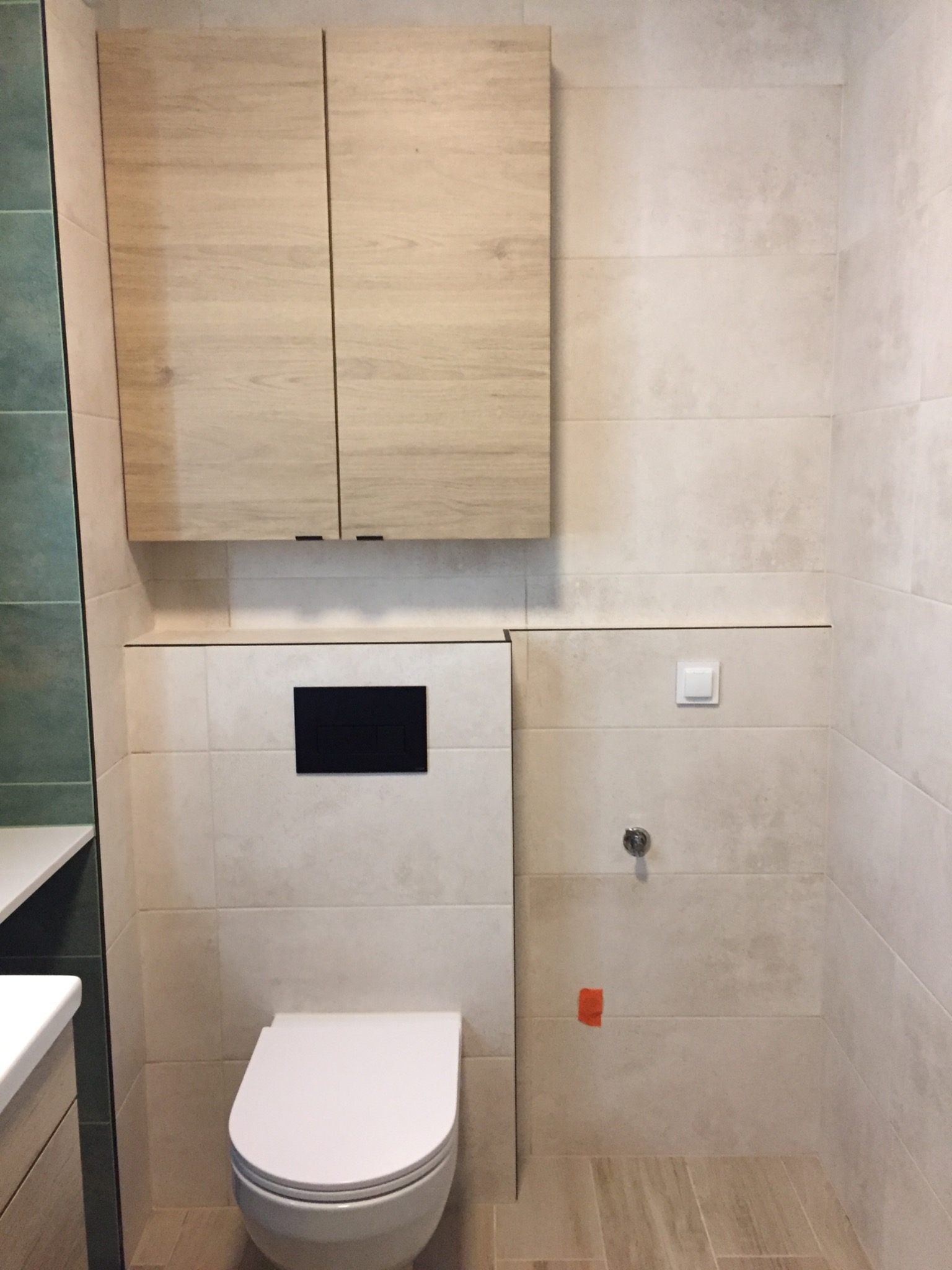 Nowoczesna łazienka z podwieszaną toaletą, szafką wiszącą z frontami imitującymi drewno i beżowymi płytkami na ścianach oraz podłodze. Widoczny czarny panel spłuczki i biały włącznik światła.