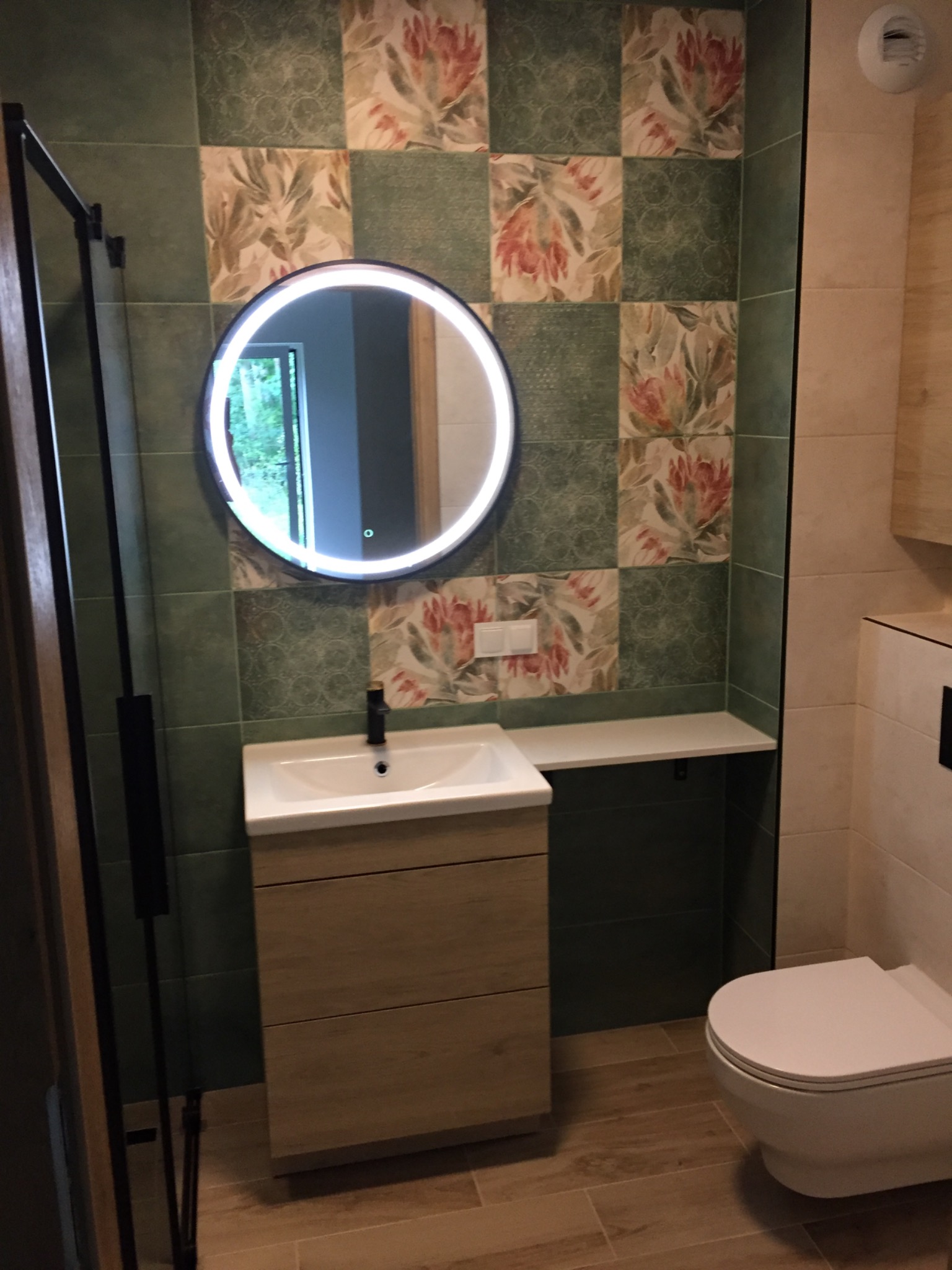 Wykończona łazienka z patchworkowymi płytkami w odcieniach zieleni i beżu z motywem kwiatowym, okrągłe lustro z podświetleniem LED nad umywalką z szafką w kolorze jasnego drewna, wisząca toaleta...