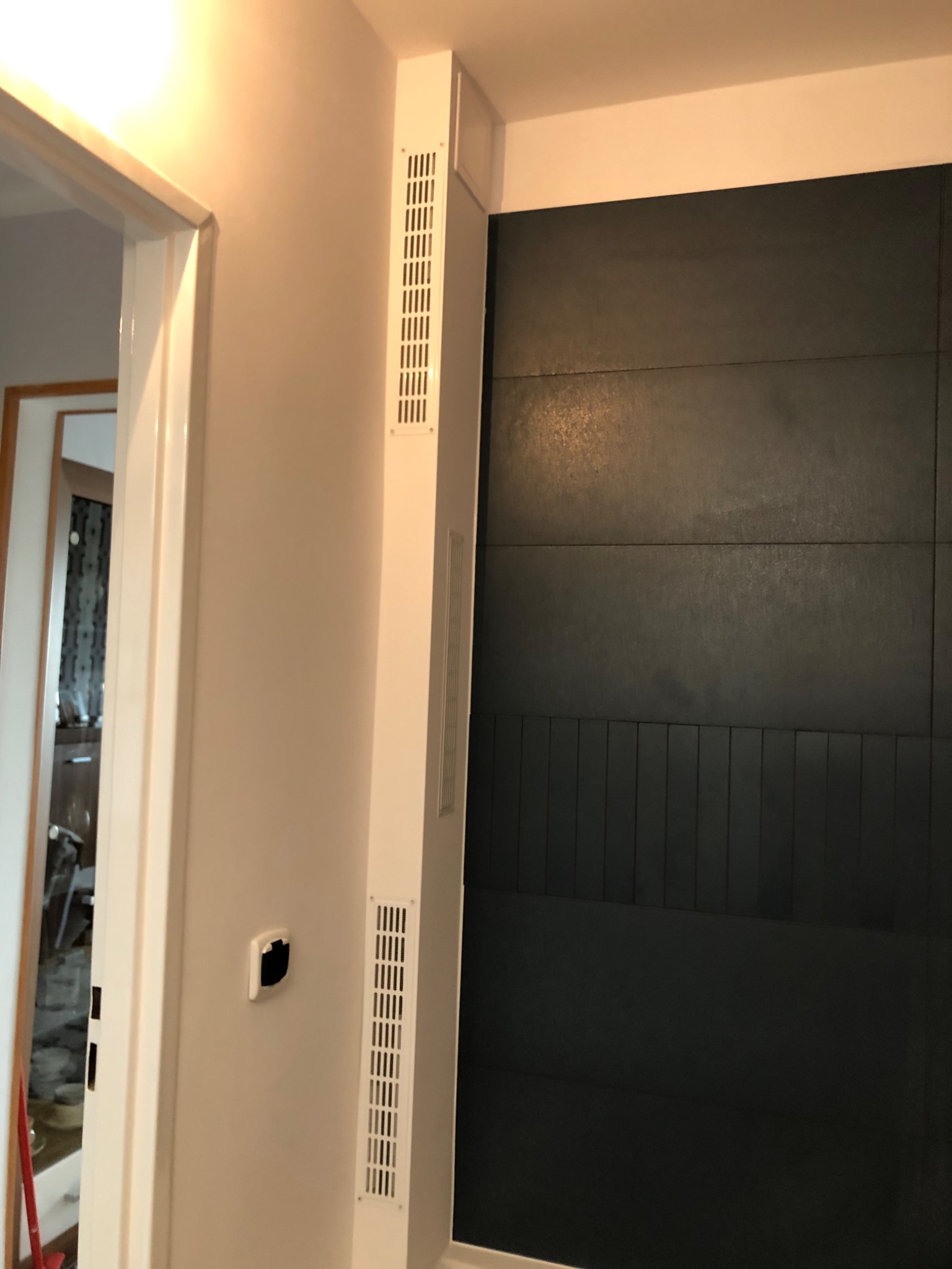 Fragment ściany z ciemnymi, poziomymi panelami dekoracyjnymi i pionowymi listwami w dolnej części, sąsiadującej z białym narożnikiem z kratkami wentylacyjnymi i otwartym przejściem do innego...