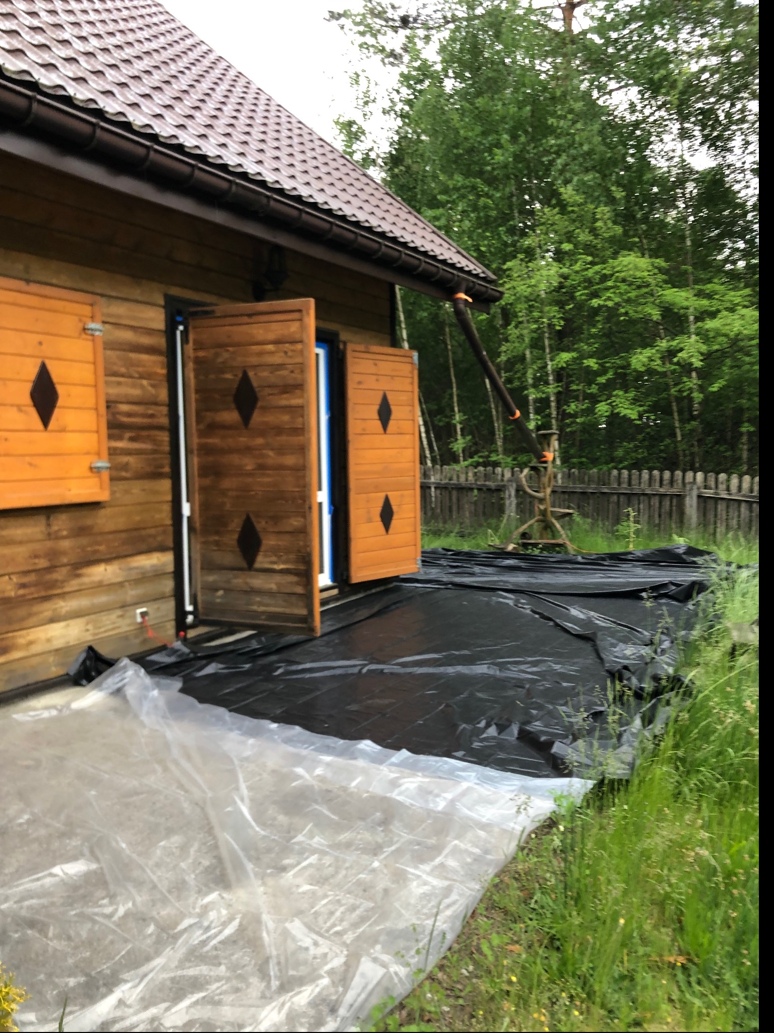 Drewniany domek z otwartymi okiennicami, zabezpieczony folią malarską przed pracami elewacyjnymi, widoczne rusztowanie w tle.