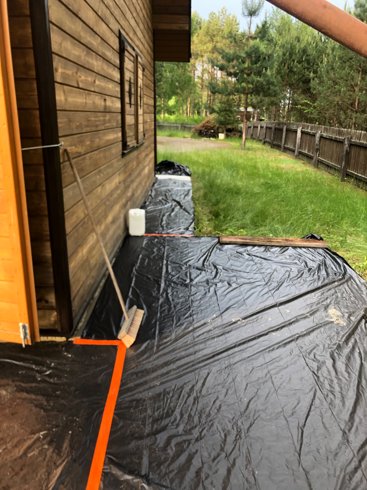 Przygotowanie do malowania drewnianej elewacji domu: zabezpieczona folią przestrzeń wokół ściany, widoczna szczotka i kanister z farbą.