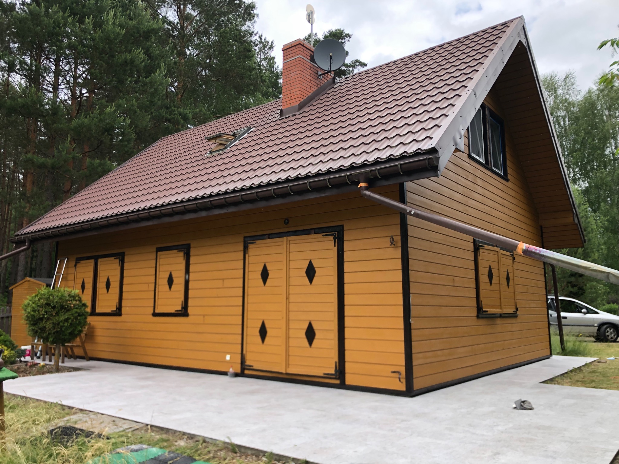 Żółty, drewniany dom z brązowym dachem z blachy trapezowej, ceglanym kominem i anteną satelitarną, widoczny z zewnątrz na tle drzew.