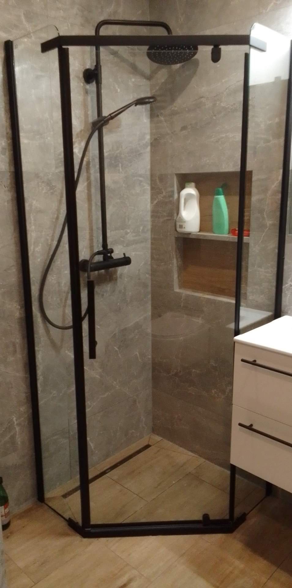 Nowoczesna kabina prysznicowa z czarnymi profilami na tle szarych płytek imitujących marmur, z wnęką na kosmetyki i drewnopodobną podłogą.