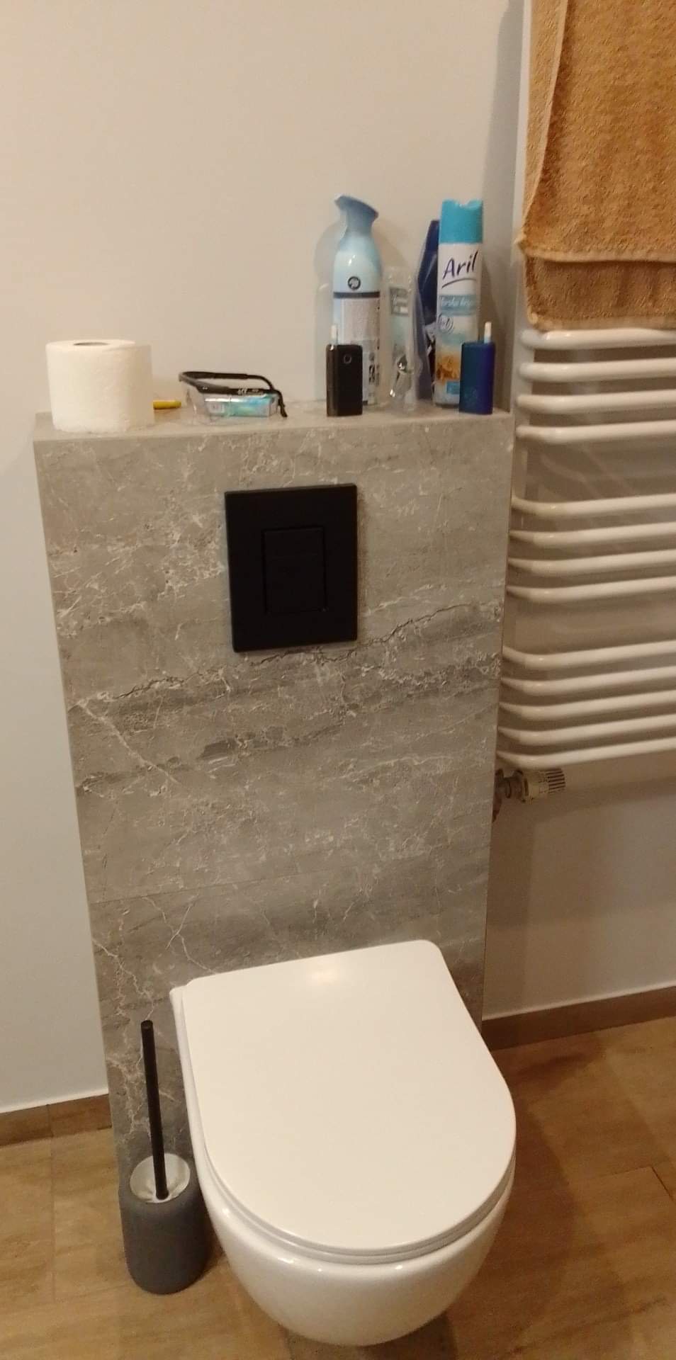 Nowoczesna toaleta wisząca z czarnym przyciskiem spłukującym na ścianie wykończonej płytkami imitującymi marmur, obok grzejnik drabinkowy i akcesoria łazienkowe.